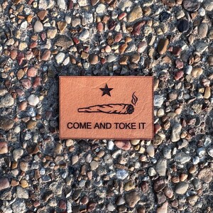 May include: A rectangular, tan leather patch with a black star and a drawing of a lit cannabis cigarette. The text "COME AND TOKE IT" is below the drawing. The patch is on a background of small, colorful stones.