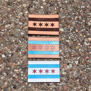 May include: Three rectangular leather patches with the Chicago city flag design. The top patch is brown with four dark stars. The middle patch has blue stripes and pink stars. The bottom patch has white and blue stripes with red stars.