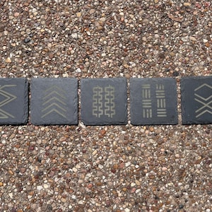 Op de afbeelding: Vijf vierkante leistenen onderzetters met geëtste geometrische ontwerpen. De onderzetters zijn donkergrijs met lichtgrijze patronen, waaronder zigzags, pijlen en parallelle lijnen. Ze zijn gerangschikt op een kiezeloppervlak.