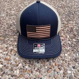 May include: A navy blue and beige trucker cap featuring a leather American flag emblem. The cap has a curved peak and an adjustable snap closure. The tag reads "RICHARDSON ORIGINAL 112 TRUCKER ADJUSTABLE".