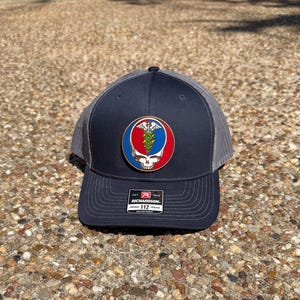 Puede incluir: Gorra de camionero azul marino con la parte trasera de malla gris y un parche redondo con una calavera, un símbolo médico y elementos botánicos en rojo, azul y verde. La gorra tiene una visera curva y el texto "RICHARDSON 112".