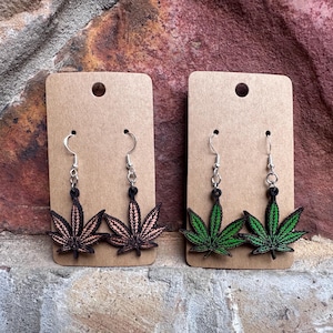 Op de afbeelding: Twee paar oorhangers met een marihuanabladontwerp. Het ene paar is zwart met roze accenten en het andere paar is groen.
