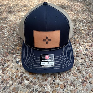 Może przedstawiać: Granatowo-beżowa czapka typu trucker z naszywką ze skóry z symbolem Zia. Czapka ma zakrzywiony daszek i regulowany pasek. Etykieta głosi "RICHARDSON ORIGINAL 112 TRUCKER ADJUSTABLE."