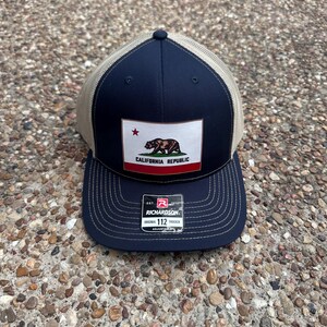 May include: Navy blue and tan trucker hat featuring the California Republic flag patch. The patch displays a bear, a star, and the words "CALIFORNIA REPUBLIC". The hat has a curved brim and an adjustable strap.