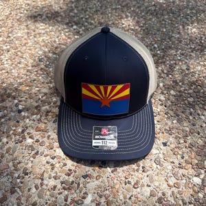 May include: A navy blue trucker hat with a tan mesh back. The front features a patch with the Arizona state flag. The hat has a curved brim and an adjustable strap. The tag reads "Richardson 112 Trucker".