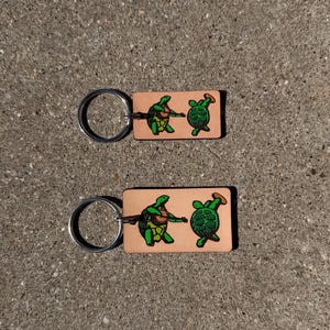 Puede incluir: Dos llaveros de cuero con fondo marrón claro. Cada llavero presenta una tortuga verde tocando la guitarra y una tortuga verde con sombrero. Los llaveros tienen un anillo de metal plateado.