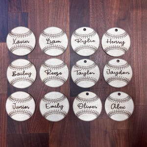 May include: Twelve wooden baseball ornaments with names engraved on them. The names are: Xavier, Liam, Rylie, Henry, Bailey, Reese, Taylor, Jaydon, Junior, Emily, Oliver, and Alex.