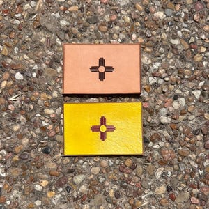 May include: Two rectangular leather patches, one peach-colored and one yellow, each featuring the Zia symbol of New Mexico. The Zia symbol is a circle with four groups of four rays extending outward. The patches are displayed on a pebbled surface.