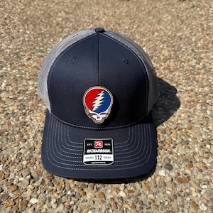 Puede incluir: Gorra de camionero azul marino con la parte trasera de malla gris y una visera curva. La gorra presenta un emblema colorido de una calavera con un diseño de rayo. La gorra es ajustable y tiene el texto "RICHARDSON" en la parte delantera.
