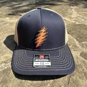 May include: A navy blue and beige trucker hat with a brown leather lightning bolt patch. The hat has an adjustable strap and a label that reads "Richardson. Est. 1970. Original 112 Trucker Adjustable."
