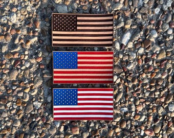 Könnte beinhalten: Drei rechteckige amerikanische Flaggen aus Holz. Die obere Flagge ist braun mit einem dunkelbraunen Sternenfeld und Streifen. Die mittleren und unteren Flaggen haben ein blaues Sternenfeld und rot-weiße Streifen. Die Flaggen liegen auf einer strukturierten Steinoberfläche.