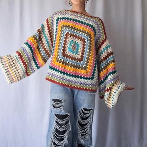 May include: A vibrant, hand-crocheted sweater featuring a square design in multiple colours, such as red, yellow, blue, and green. It has long, flared sleeves and a wide neckline. The sweater is worn with light blue ripped jeans.