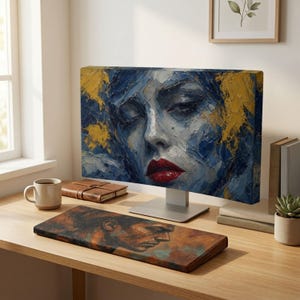 Könnte beinhalten: Ein Computermonitor zeigt ein Ölgemälde eines Gesichts in Blau- und Goldtönen mit leuchtend roten Lippen. Unter dem Monitor liegt eine rechteckige Handgelenkauflage mit einem braun-orangen Porträt. Eine Tasse und Bücher stehen daneben.