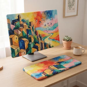 Könnte beinhalten: Eine Computermonitor-Abdeckung und eine passende rechteckige Schreibtischunterlage mit einer lebendigen Aquarellillustration eines Dorfes am Hang mit bunten Häusern, einem gewundenen Pfad und Vögeln vor einem orangefarbenen und blauen Himmel.