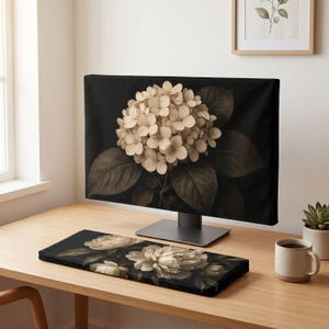 May include: A computer monitor displaying a large beige hydrangea flower on a black background, paired with a matching rectangular keyboard wrist rest featuring a floral design of white peonies on a black background, set on a wooden desk.