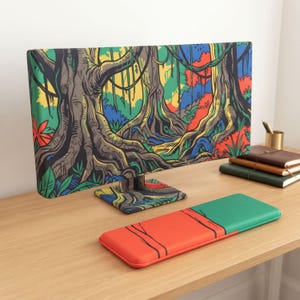Puede incluir: Una funda para monitor de computadora y un reposamuñecas para teclado a juego. La funda del monitor presenta una vibrante ilustración de selva con grandes troncos de árboles, enredaderas y follaje colorido. El reposamuñecas está dividido en secciones roja y verde.