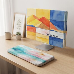 May include: A computer monitor cover featuring a watercolor desert landscape with yellow and orange dunes and a line of four small black antelopes, paired with a matching keyboard wrist rest displaying a soft blue, pink, and white watercolor wash.