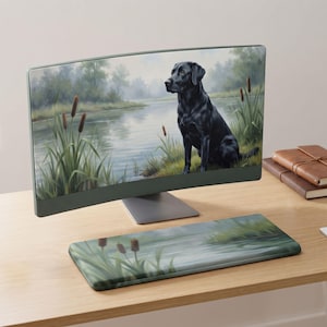 Puede incluir: Un monitor de computadora y un reposamuñecas para teclado a juego con un paisaje impreso de un labrador negro sentado a la orilla de un río con juncos verdes y espadañas marrones bajo un cielo suave y nublado.