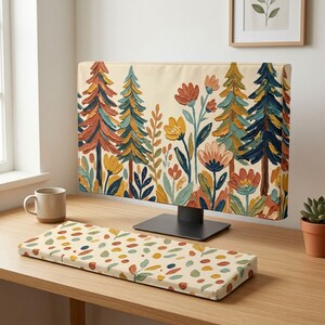 Op de afbeelding: Een computermonitorhoes en een bijpassende toetsenbordpolssteun met een olieverfschilderij-stijl print van dennenbomen en diverse bloemen in tinten oranje, geel, blauw en groen op een crèmekleurige achtergrond, geplaatst op een houten bureau.