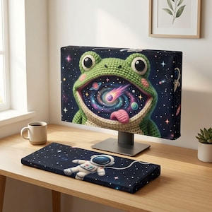 May include: A computer monitor cover and matching keyboard wrist rest featuring a crochet-style green frog with a wide open mouth revealing a colourful galaxy, stars, and a comet, set against a dark blue space background with a floating astronaut.