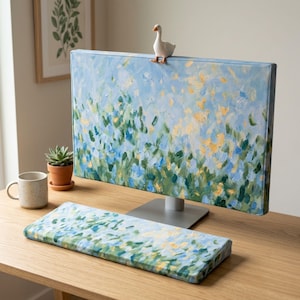 Puede incluir: Una funda para monitor de ordenador y un reposamuñecas a juego para teclado con un patrón pintado impresionista de pinceladas azules, verdes y amarillas. Una pequeña figura de ganso de cerámica blanca se asienta sobre el borde superior de la funda.