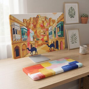 Pode incluir: Uma capa para monitor de computador com uma ilustração colorida de uma fortaleza no deserto com varandas em arco e dois camelos azul-escuros em primeiro plano, acompanhada por um apoio de pulso retangular para teclado com um padrão de pintura abstrata multicolorida.