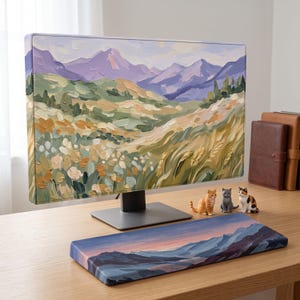May include: A computer monitor displaying a landscape painting with purple mountains and green fields, accompanied by a matching rectangular wrist rest featuring a sunset mountain scene, and three small cat figurines on the desk surface.
