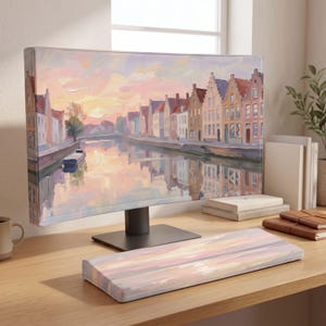 May include: A computer monitor and a matching keyboard wrist rest featuring an oil painting style print of a canal-side town at sunset. The scene depicts rows of buildings with stepped gables reflecting in the water under a soft pink and orange sky.