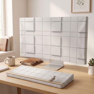 Puede incluir: Una funda blanca para monitor de computadora y un reposamuñecas para teclado a juego, ambos con un patrón de cuadrícula de líneas grises finas. La funda del monitor tiene paneles cuadrados elevados que crean una apariencia geométrica texturizada.