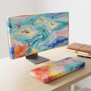 Könnte beinhalten: Ein Computermonitor und eine rechteckige Handgelenkauflage mit Aquarell-Design. Der Monitor zeigt eine Insel-Szene mit blauem Wasser und Vögeln, während die Handgelenkauflage ein weiches, abstraktes Muster in Rosa, Gelb, Orange und Blau aufweist.