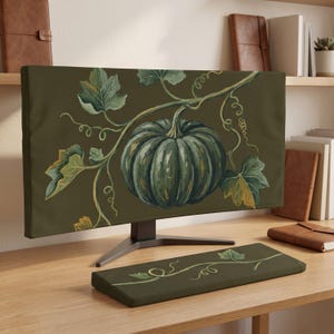 Op de afbeelding: Een computermonitorhoes en bijpassende toetsenbordpolssteun met een donker olijfgroene achtergrond en een centrale illustratie van een donkergroene pompoen aan een rank met grote bladeren en krullende ranken.