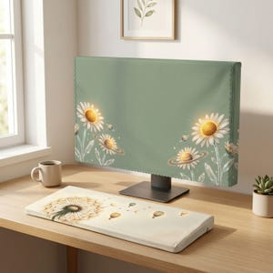 May include: A sage green computer monitor cover featuring white daisy flowers with glowing yellow centres and small planetary rings, paired with a rectangular off-white keyboard wrist rest decorated with a dandelion seed head and hot air balloons.