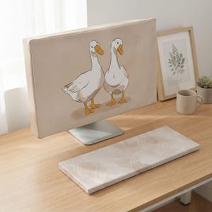 Könnte beinhalten: Eine beige Computer-Monitorabdeckung mit zwei weißen Enten mit orangefarbenen Schnäbeln und Füßen, kombiniert mit einer passenden rechteckigen Tastatur-Handballenauflage mit weißem Federmuster auf beigem Grund.