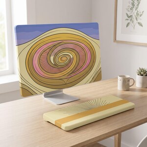 May include: A computer monitor displaying a graphic of a golden spiral pattern with pink accents against a blue sky background, accompanied by a matching rectangular keyboard wrist rest featuring a sunburst design in yellow and tan tones.
