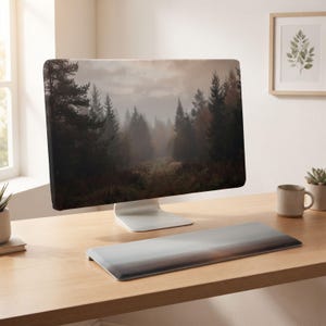 Könnte beinhalten: Ein Computermonitor mit einem Bildschirmfoto einer nebligen Waldlandschaft mit hohen Kiefern und einem Feldweg. Der Monitor hat einen dünnen silbernen Standfuß und steht auf einem Holztisch neben einer passenden Tastatur-Handballenauflage.