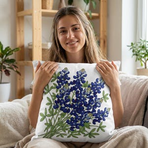 Op de afbeelding: Vierkant wit kussen met een gedetailleerde illustratie van lupinebloemen. Het ontwerp toont een cluster van levendige blauwe bloemen met groene bladeren en stelen tegen een witte achtergrond. Een decoratief accent voor een woon- of slaapkamer.
