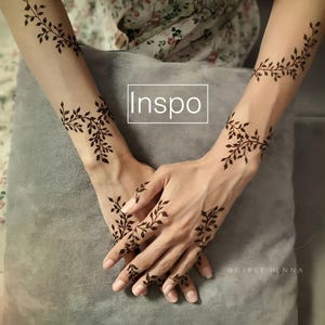 May include: Henna art featuring a delicate leaf design on the wrists, forearms, and fingers. The dark brown henna is applied in a vine-like pattern, with the word "Inspo" in a white box. The image is taken from a close-up angle.