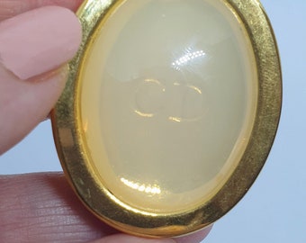 May include: Oval-shaped gold-tone button with a cream-colored center. The center features the embossed initials "CD". The button has a smooth, glossy finish and is held by a hand.