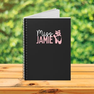 May include: A black spiral-bound notebook featuring the white text Miss and the pink text JAMIE, accompanied by a graphic of pink ballet slippers. The notebook has a clean, matte cover design with a professional wire-o binding along the left side.
