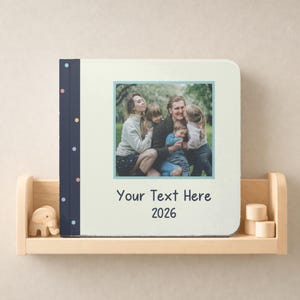 May include: A square photo book with a cream-colored cover featuring a central square photograph of a family, a dark blue spine with colorful dots, and the printed text "Your Text Here" above the year "2026" in a simple dark font.