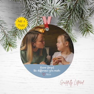 May include: A round, silver ornament with a photo of two people. The ornament has a red ribbon for hanging and a blue section with the text "Never forget the difference you make" and the name "Alissa". The words "Add a Photo!" are in a yellow starburst.
