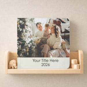 May include: A square photo book with rounded corners featuring a family portrait in front of a Christmas tree. The cover includes the printed text Your Title Here 2026. The book is displayed on a light-colored wooden shelf with small wooden toys.