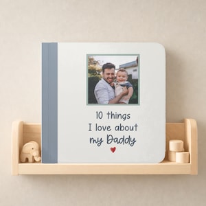 May include: A square board book with a white cover featuring a blue spine and a centered photograph of an adult holding a baby. The text reads 10 things I love about my Daddy with a small red heart icon below the text on the front cover.