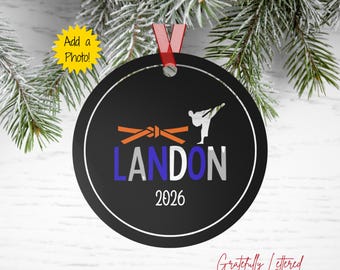 May include: A round black holiday ornament featuring a white silhouette of a person performing a karate kick, an orange martial arts belt, the name LANDON in blue and white letters, and the year 2026 in white, suspended by a red ribbon.