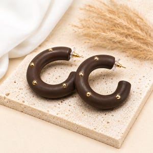 May include: A pair of dark brown hoop earrings with gold-coloured stud accents. The earrings are C-shaped and have a smooth, matte finish. The earrings are displayed on a textured, light-coloured surface.