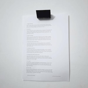 Könnte beinhalten: Eine schwarze, rechteckige Büroklammer hält ein weißes Blatt Papier vor einem weißen Hintergrund. Das Papier enthält gedruckten Text, wahrscheinlich eine Liste oder ein Dokument. Die Klammer ist oben zentriert.