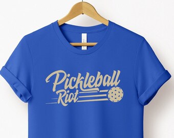 May include: A vibrant blue t-shirt featuring the text "Pickleball Riot" in a tan, cursive font. The design incorporates a pickleball and speed lines. The tee is displayed on a wooden hanger.