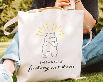May include: White tote bag with a black line drawing of a cat with its arms crossed. The cat is surrounded by yellow sun rays. The text "I am a ray of fucking sunshine" is printed in black below the cat.