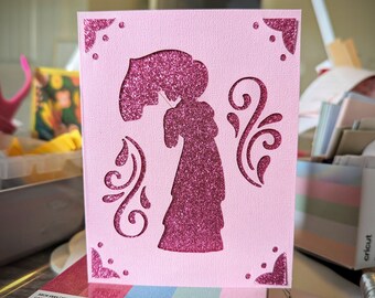 May include: A pink greeting card with a glittery silhouette of a woman holding a parasol. The card features decorative corner accents and swirling cut-out designs. The woman's silhouette is filled with sparkling pink glitter.