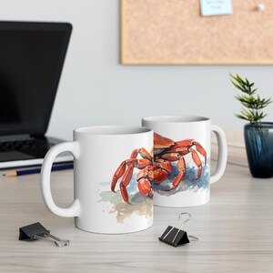May include: Two white ceramic mugs with a watercolor illustration of a crab. The crab is depicted in shades of red and orange, with blue and tan accents. The mugs are on a light-colored wooden surface.